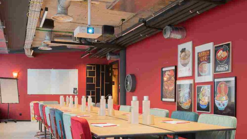 A meeting room with red walls, a long pale table lined with red and sea-green vintage chairs, under a wooden ceiling with exposed ducting.