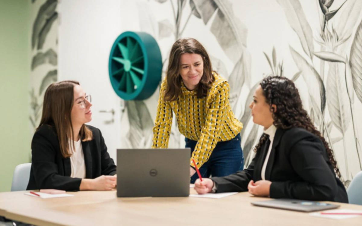 Three women talk around a laptop in a meeting room decorated with a botanical wall mural.