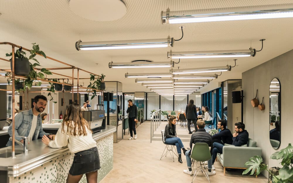 People chatting at the counter and in armchairs in a long Comet common area lit by tube lights and dotted with hanging plants.