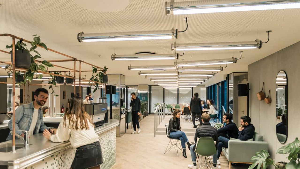 People chatting at the counter and in armchairs in a long Comet common area lit by tube lights and dotted with hanging plants.