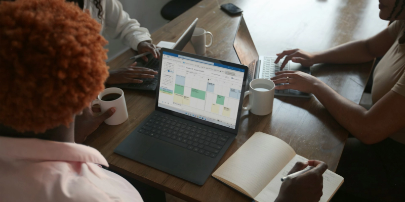 Group of female colleagues at a desk with a laptop showing Outlook Calendar