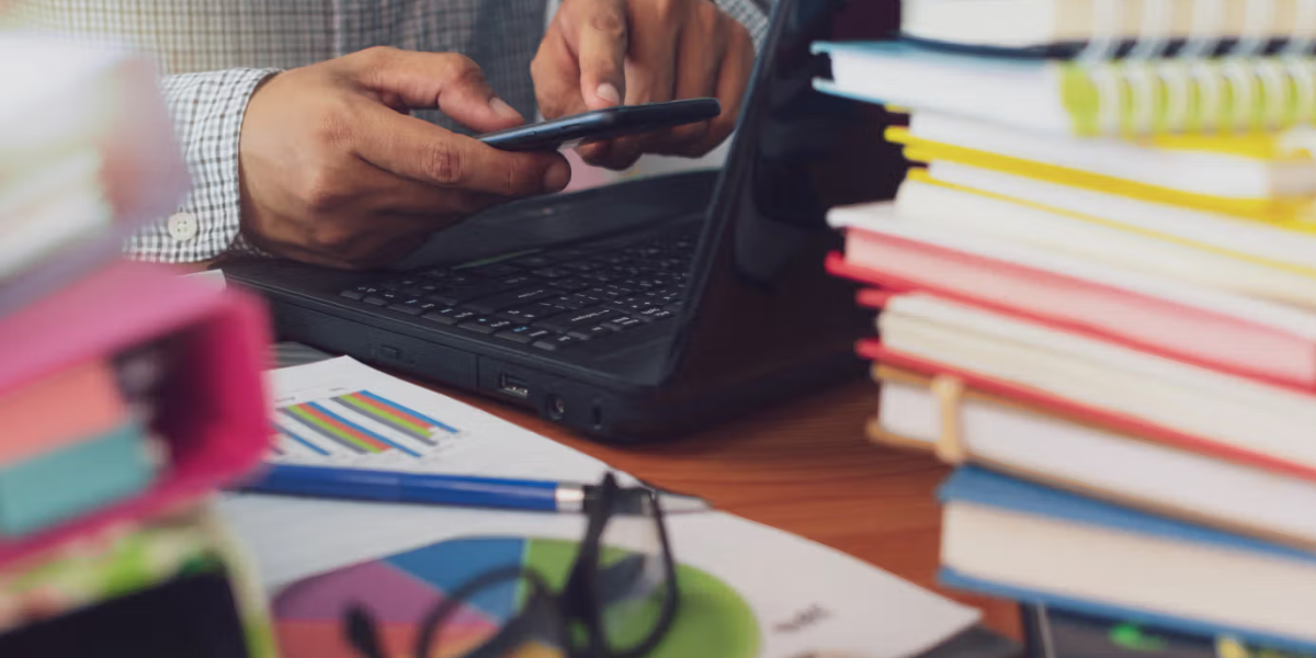 Teacher reviewing reports on a laptop and phone amid books, highlighting the need for school operations software.