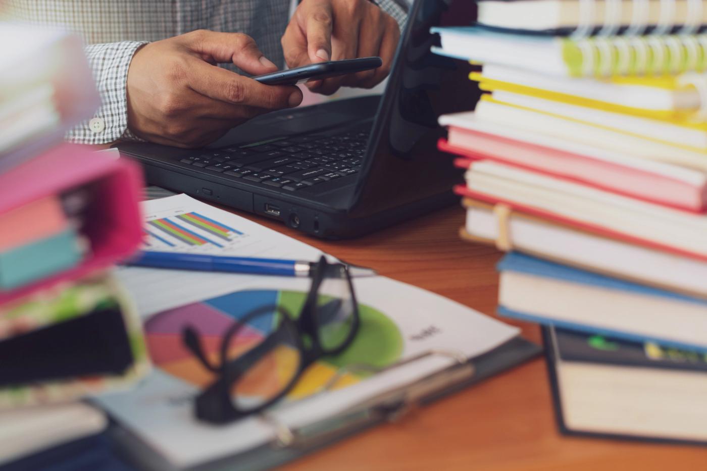 Teacher reviewing reports on a laptop and phone amid books, highlighting the need for school operations software.