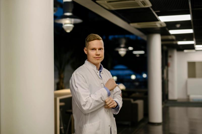 A man dressed in a white lab coat stands confidently in a modern, well-lit lobby area, adjusting his sleeve.