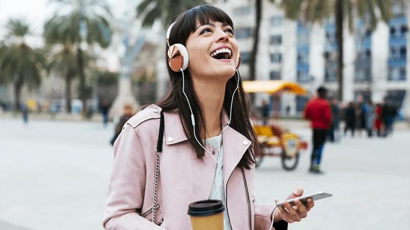 A laughing woman in a beach boulevard, listening to music from her mobile phone and headphones