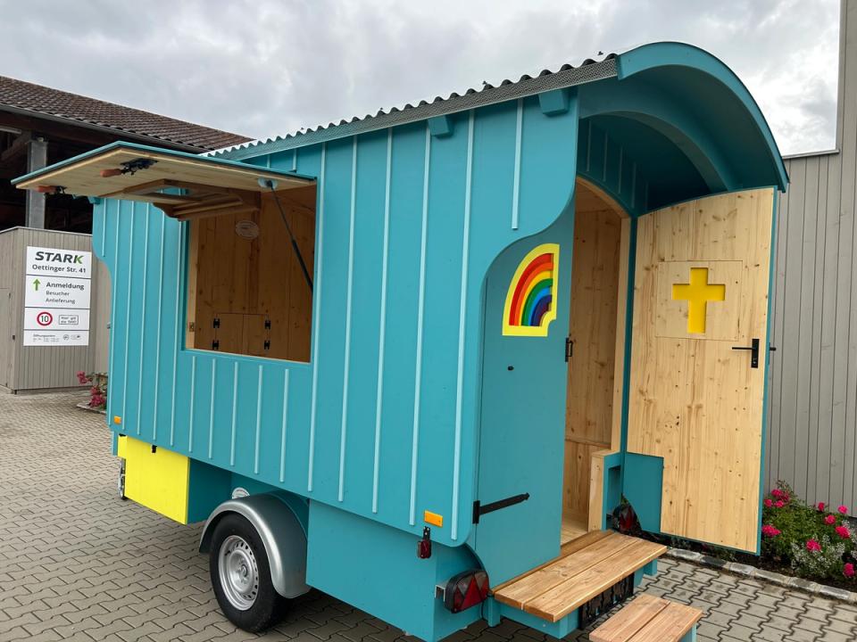 Türkiser Kirchenwagen mit aufgeklappter Seitenwand, offener Holztür mit gelbem Kreuz und Trittstufen
