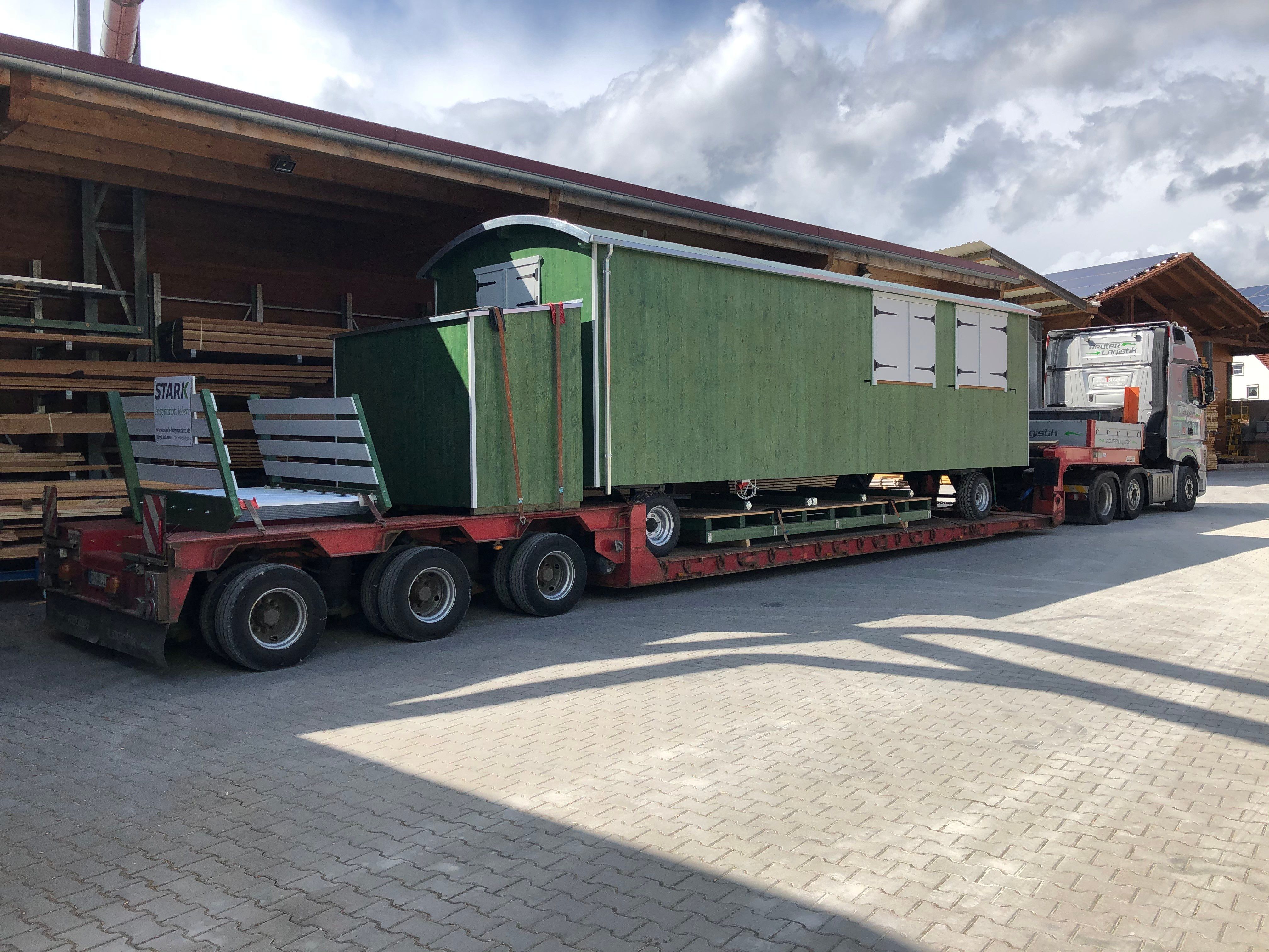 Grüner Waldkindergartenwagen auf einem Tieflader vor dem Holzlager der Zimmerei, bereit zur Auslieferung