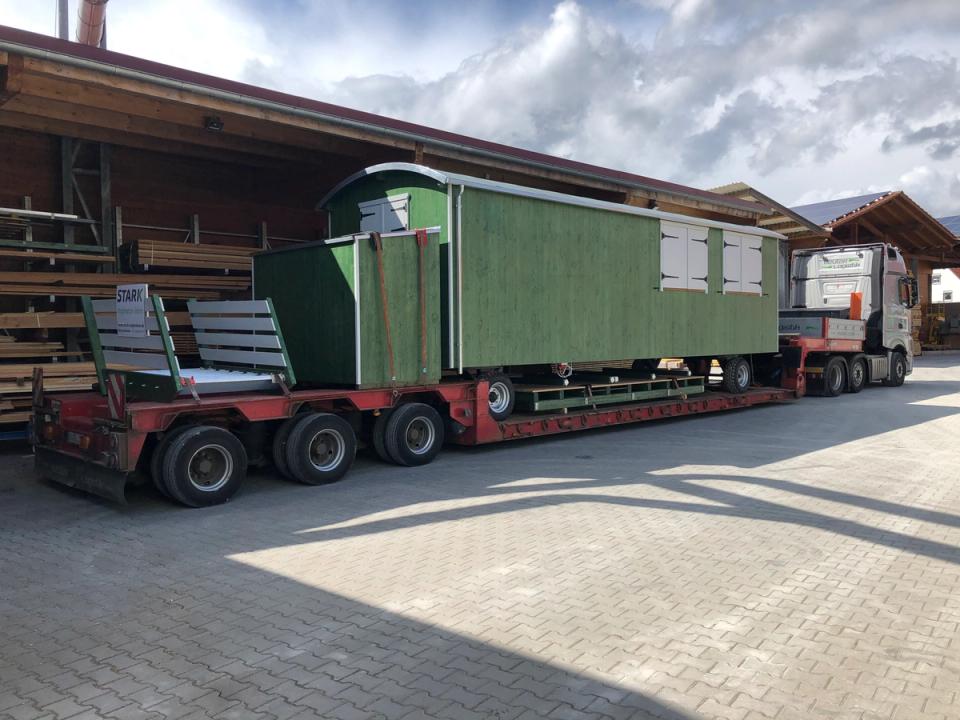 Grüner Waldkindergartenwagen auf einem Tieflader vor dem Holzlager der Zimmerei, bereit zur Auslieferung