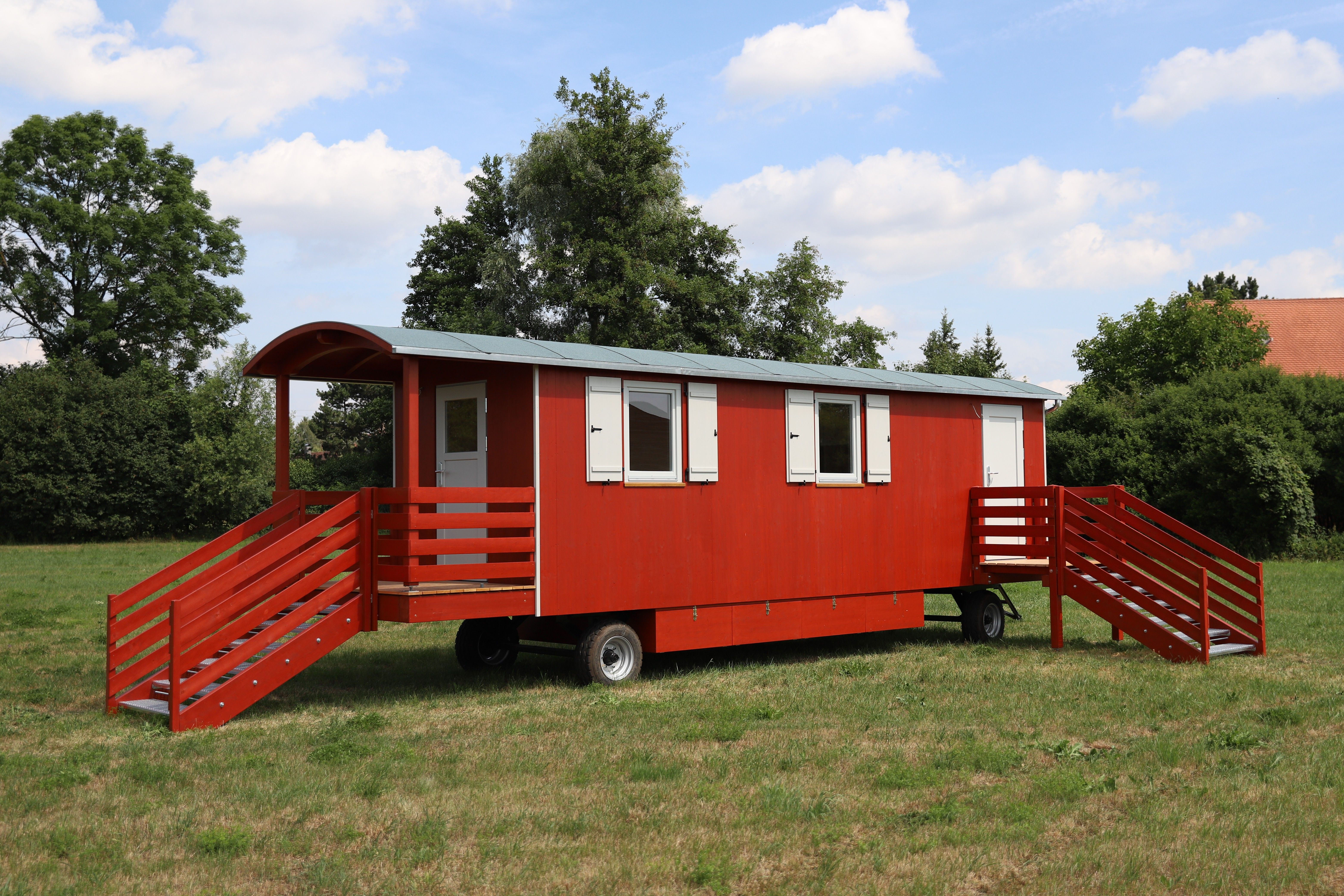 Roter Waldkindergartenwagen mit weißen Fensterläden, überdachter Veranda und zwei Treppen, von vorn auf der Wiese