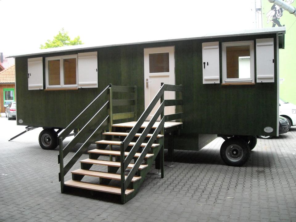 Dunkelgrüner Waldkindergartenwagen mit weißen Fensterläden und Holztreppe im Hof der Zimmerei