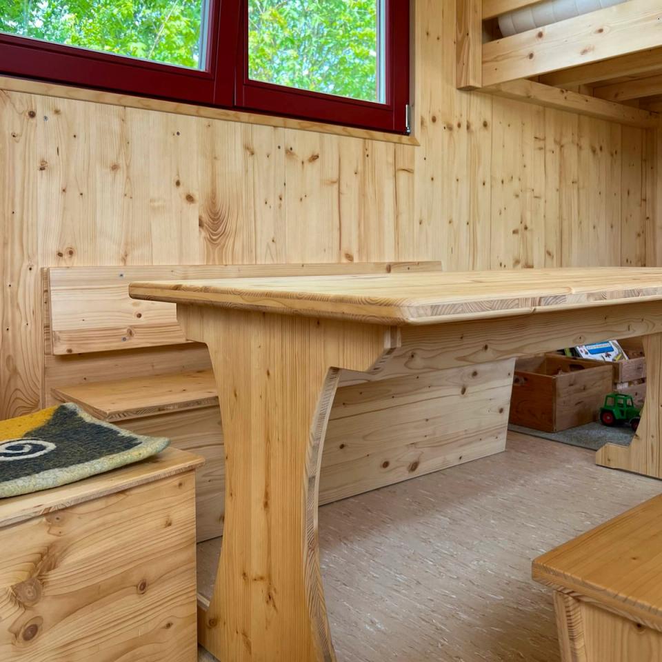 Tisch und Bank aus hellem Holz unter einem Fenster mit rotem Rahmen im Waldkindergartenwagen