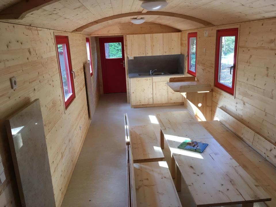Innenraum eines Waldkindergartenwagens mit Tischen, Bänken, Küchenzeile und roten Fensterrahmen