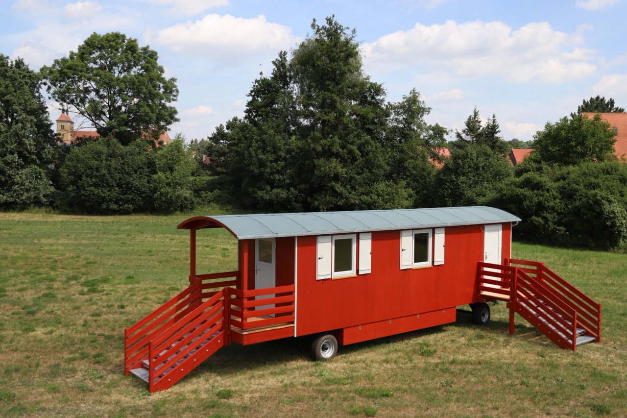 Roter Waldkindergartenwagen mit gewölbtem Dach, Veranda und Treppen an beiden Enden auf einer Wiese, dahinter Bäume und ein Kirchturm