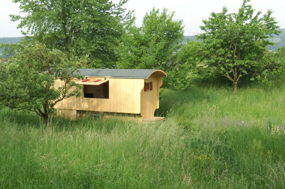 Schäferwagen aus hellem Holz im hohen Gras zwischen Obstbäumen