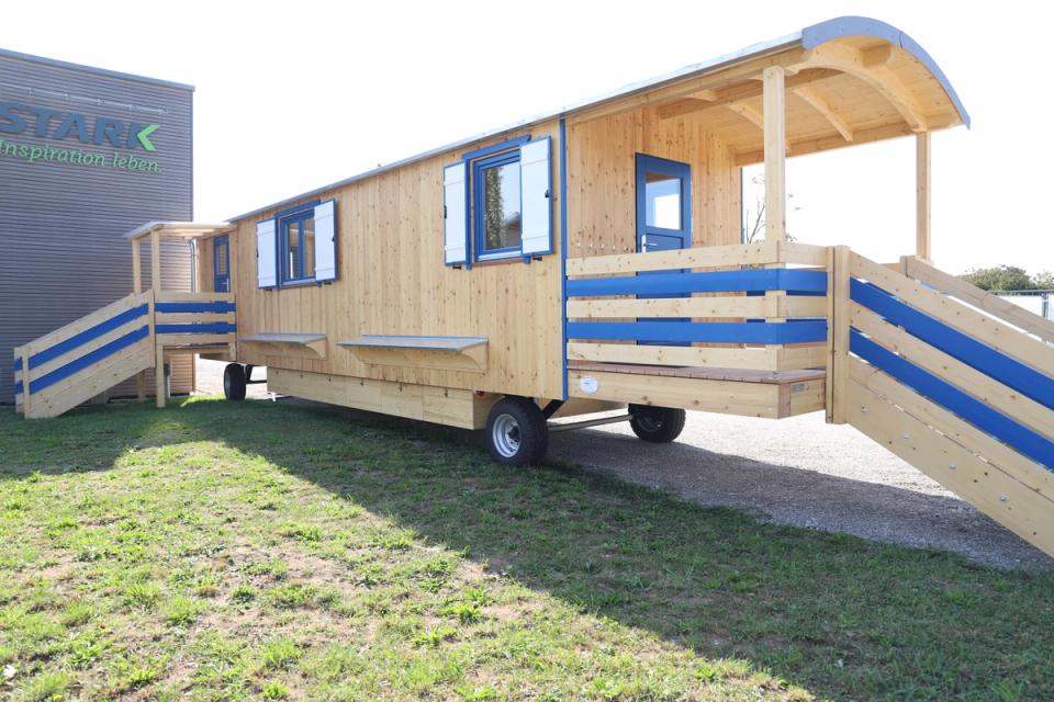 Langer Waldkindergartenwagen aus hellem Holz mit überdachter Veranda und blau gestreiften Geländern auf dem Rasen vor der Halle