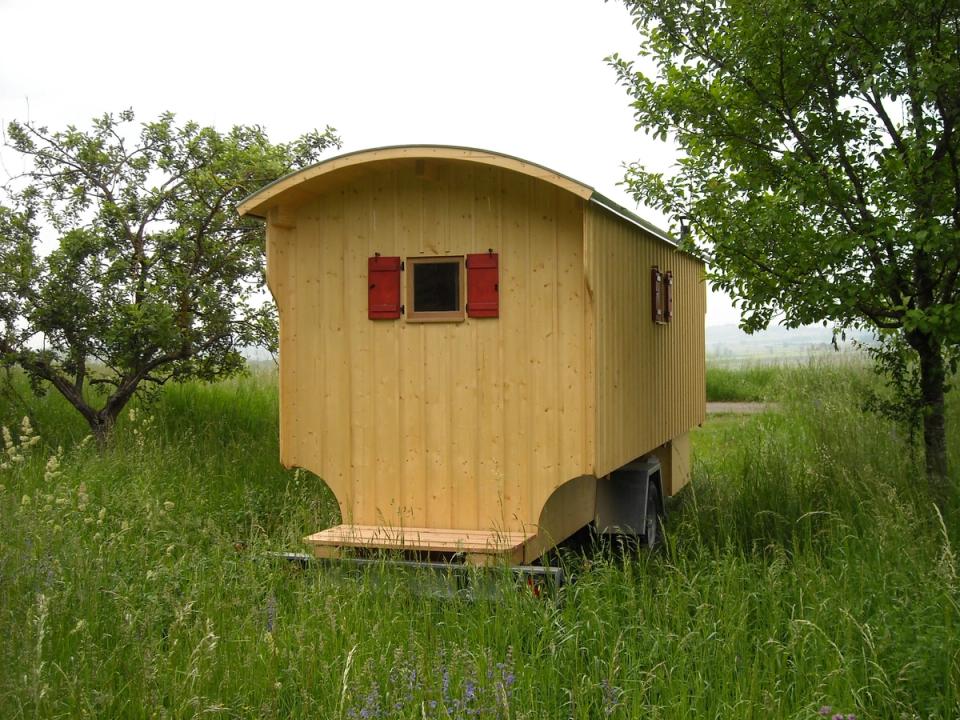 Rückseite eines Schäferwagens aus hellem Holz mit rotem Fensterladen auf einer Wiese