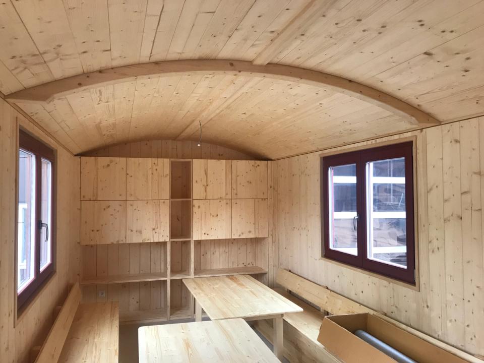 Innenraum aus hellem Holz mit Tischen, Bänken und Regalwand unter dem gewölbten Dach