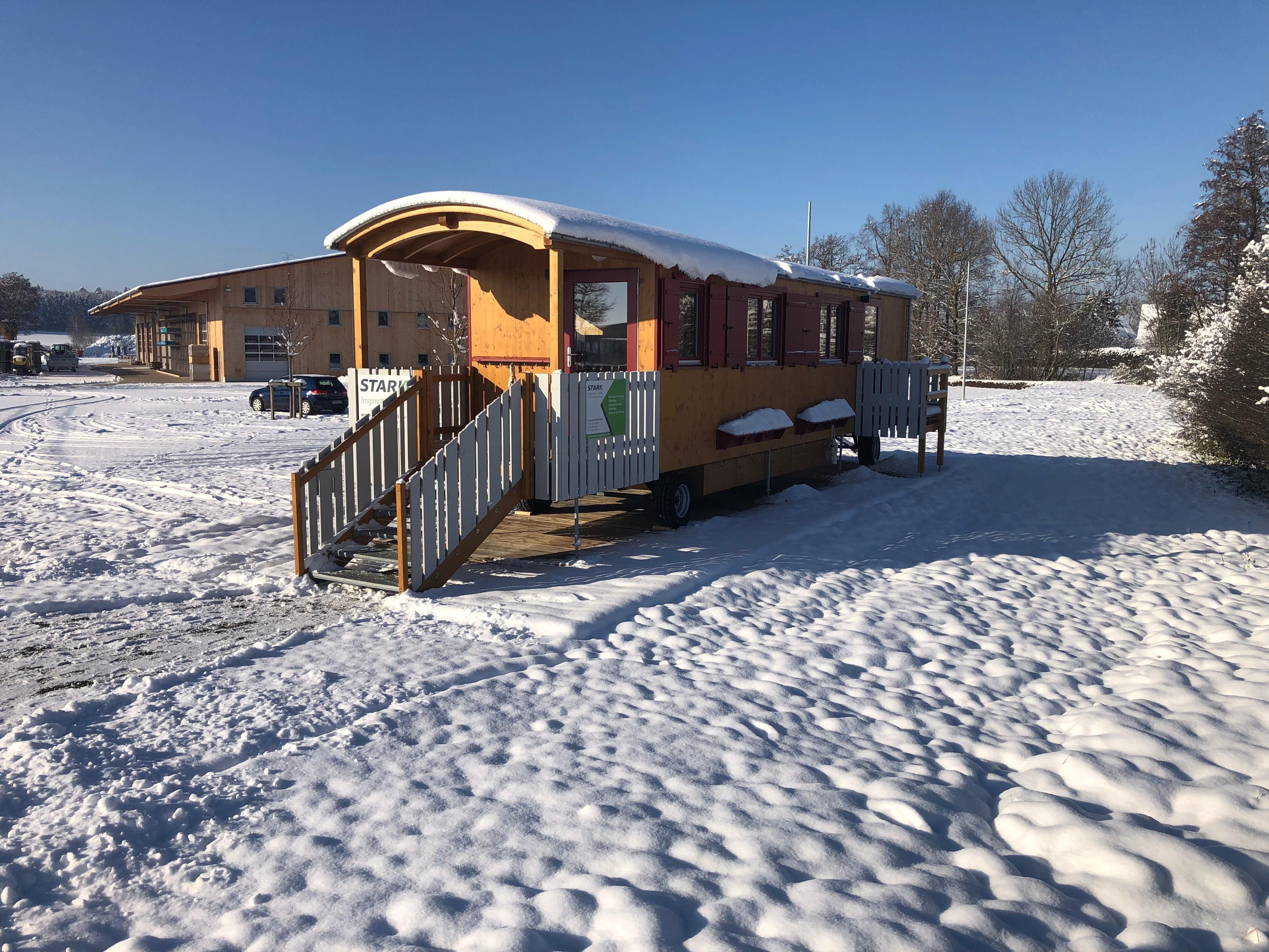 Waldkindergartenwagen mit Veranda und weißem Geländer vor der Halle der Zimmerei im Schnee