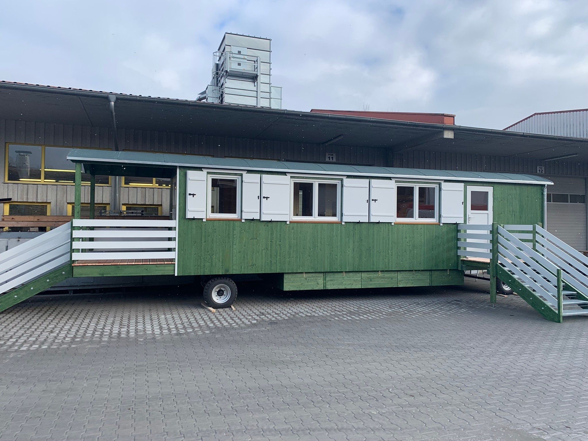 Langer grüner Waldkindergartenwagen mit weißen Fensterläden, überdachtem Eingang und Treppen an beiden Enden