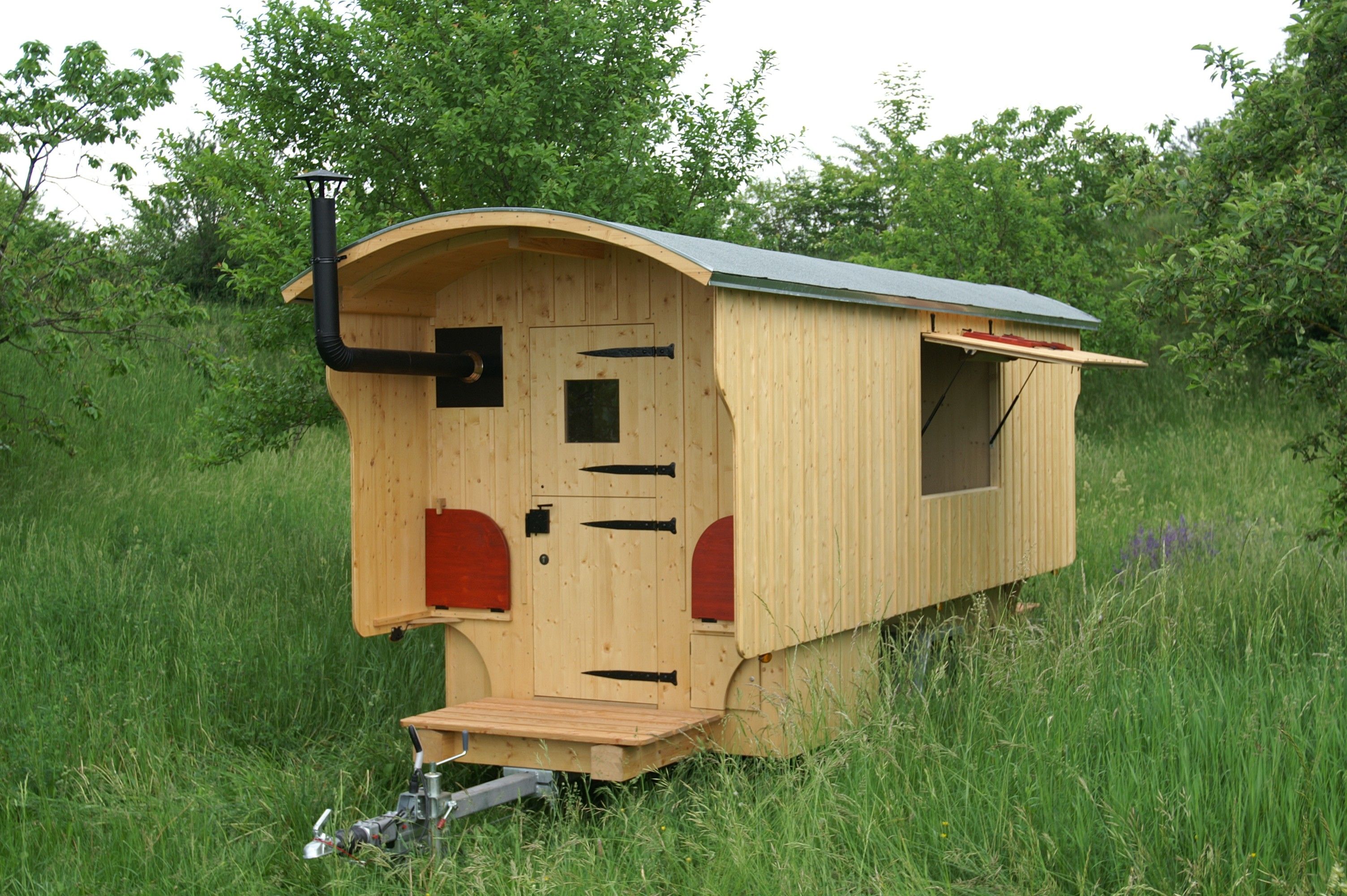 Schäferwagen aus hellem Holz mit Ofenrohr und aufgeklapptem Seitenfenster im hohen Gras