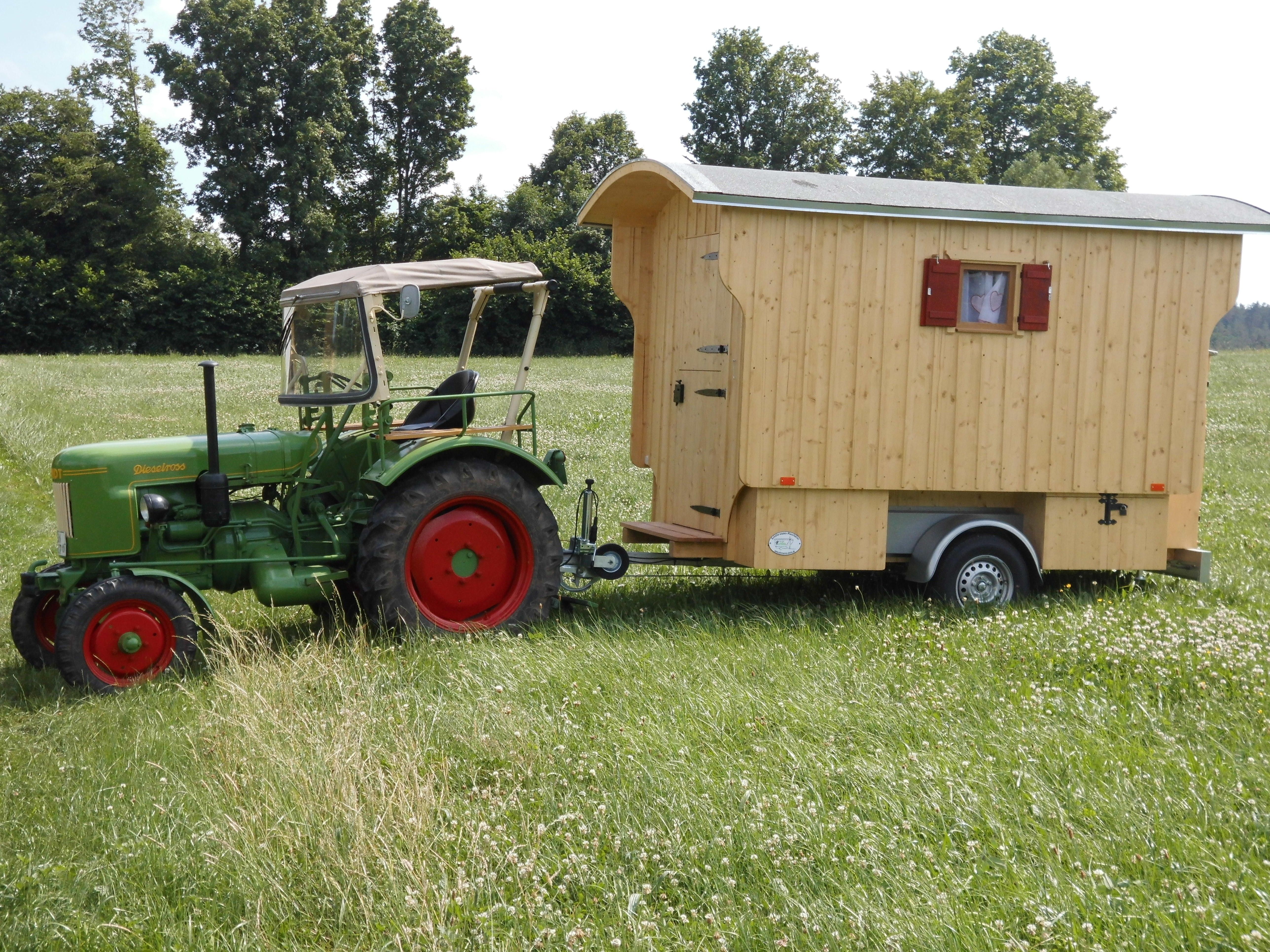 Grüner Oldtimer-Traktor mit angehängtem Schäferwagen aus hellem Holz auf einer Wiese