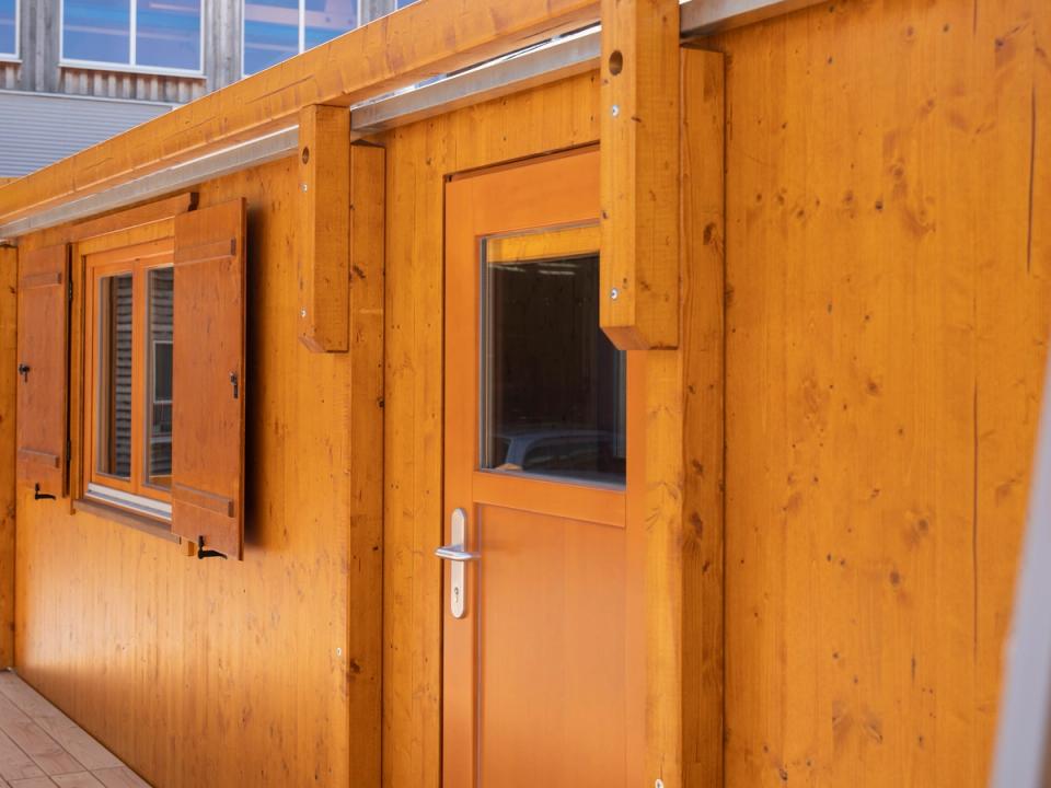 Eingangstür und Fenster mit Läden an einem honigfarbenen Waldkindergartenwagen, davor die Terrasse