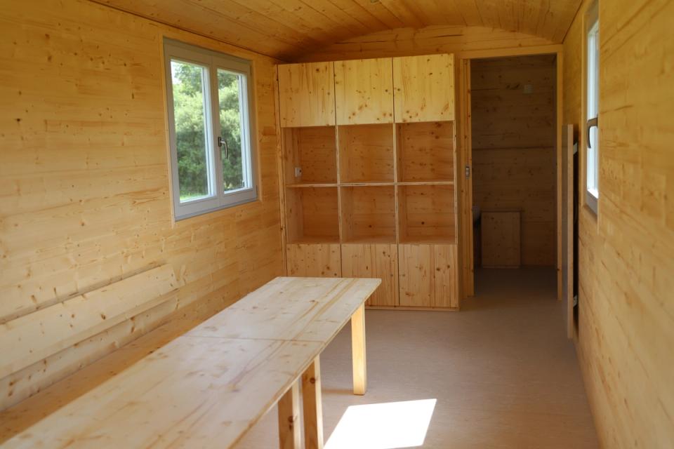 Innenraum eines Waldkindergartenwagens mit langer Bank, Regalwand mit Fächern und offener Tür zum Nebenraum