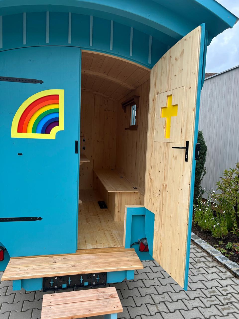 Offene Tür des Kirchenwagens mit Regenbogen und Kreuz, Blick auf die Sitzbank im Innern