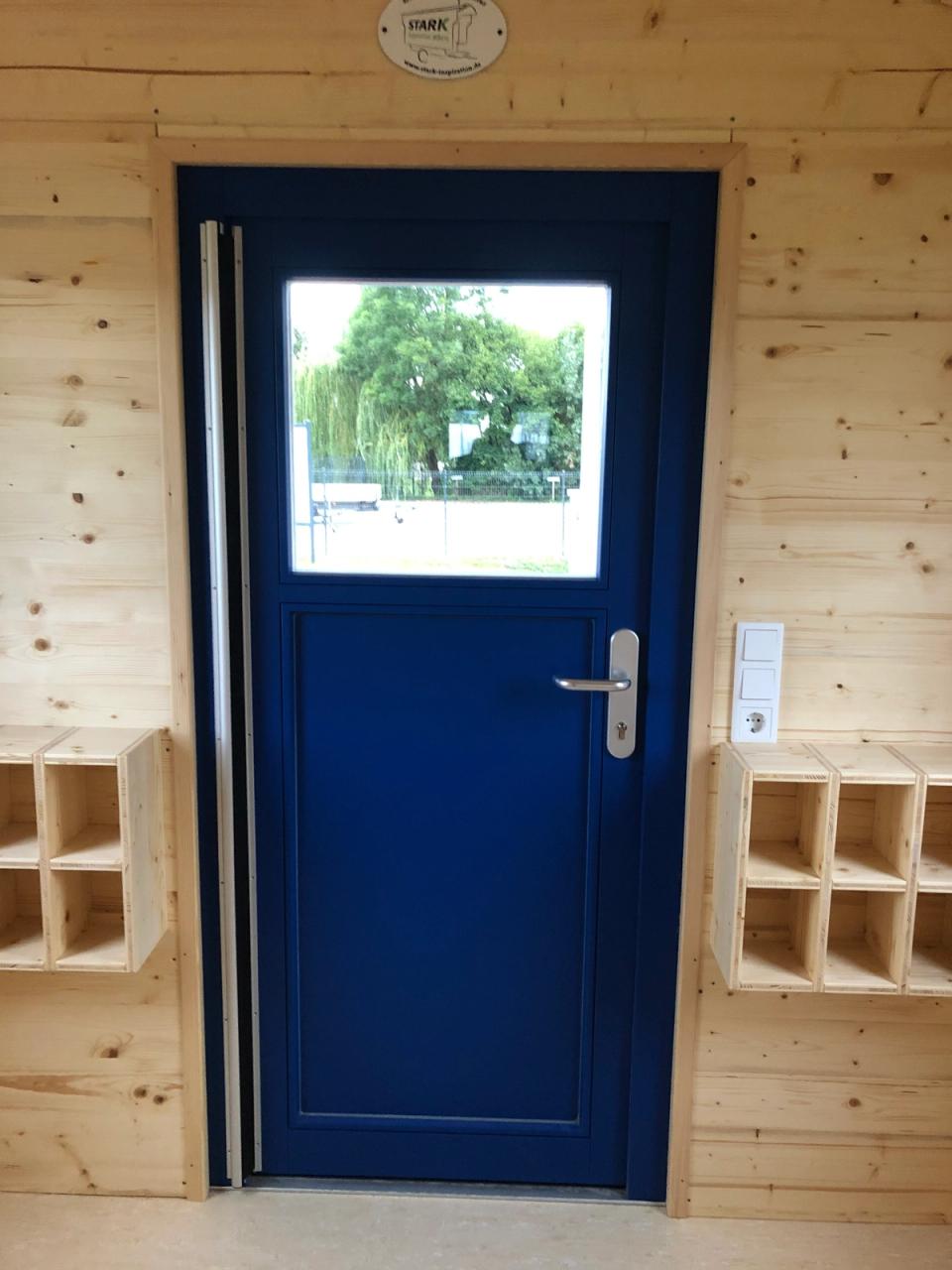 Blaue Eingangstür mit Fenster, links und rechts Fächer für Schuhe, von innen gesehen