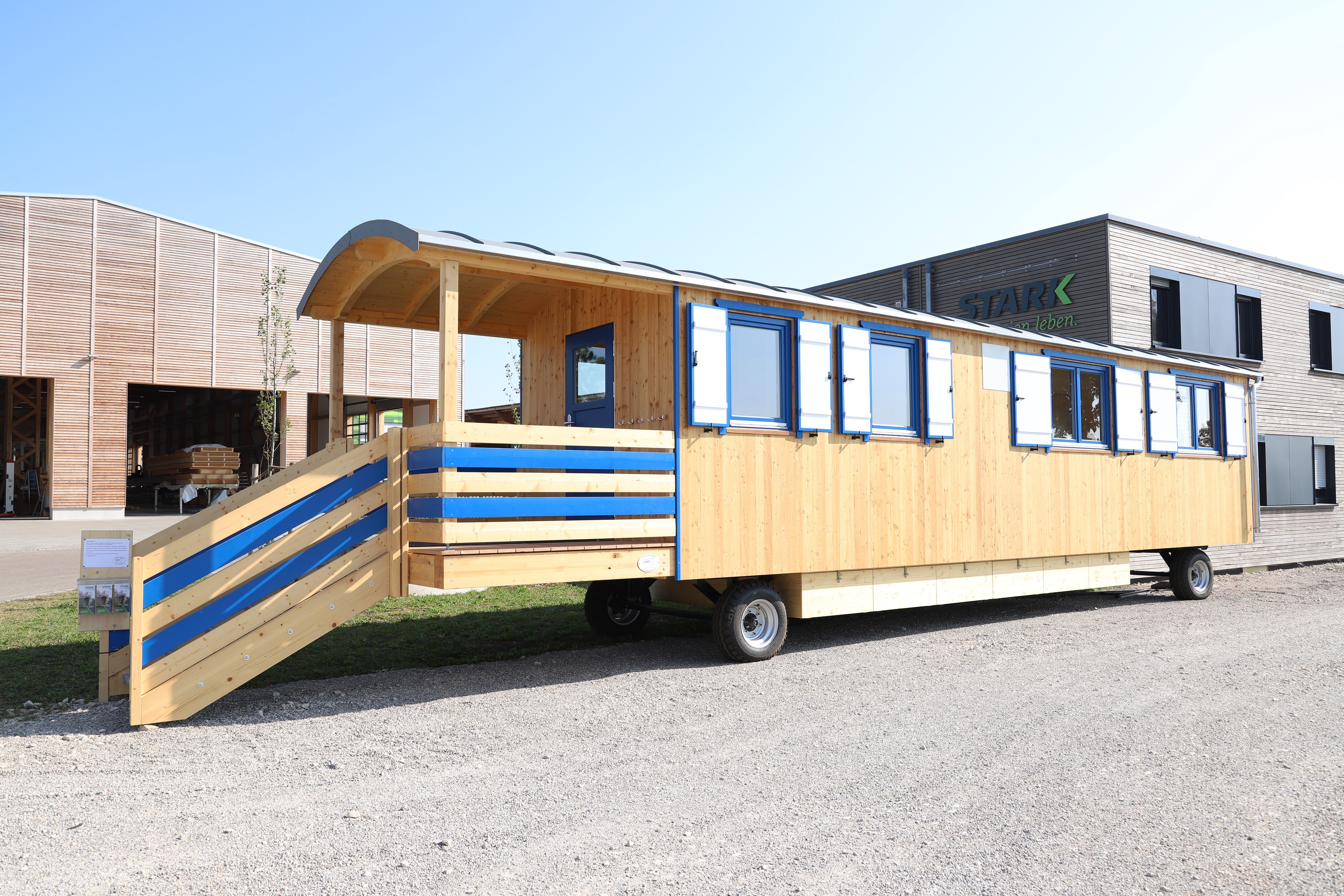Langer Waldkindergartenwagen aus hellem Holz mit blau gestreiftem Geländer, Veranda und Rampe vor der STARK-Halle