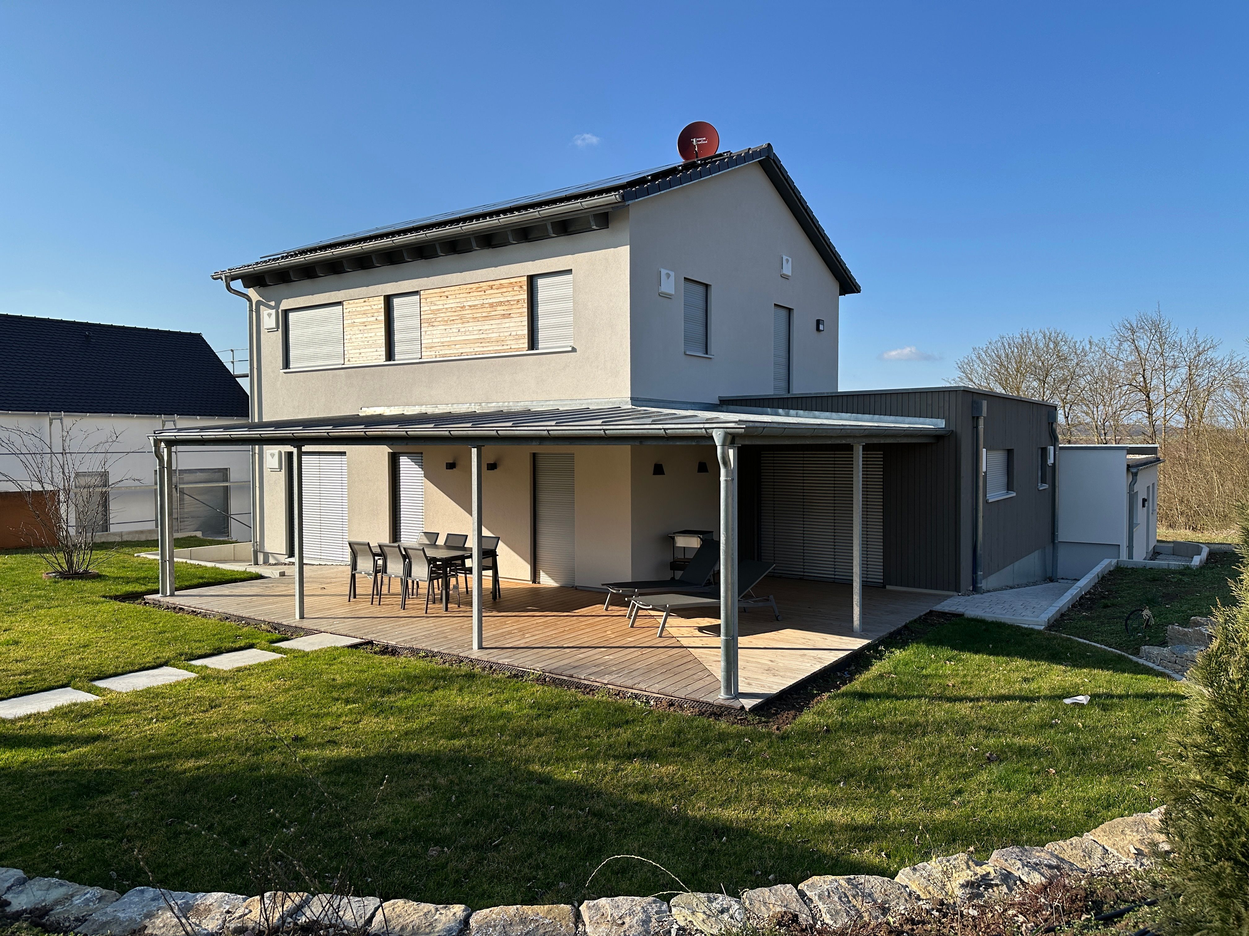 Rückseite des Wohnhauses mit Metall-Pergola über der Holzterrasse und Gartenmöbeln