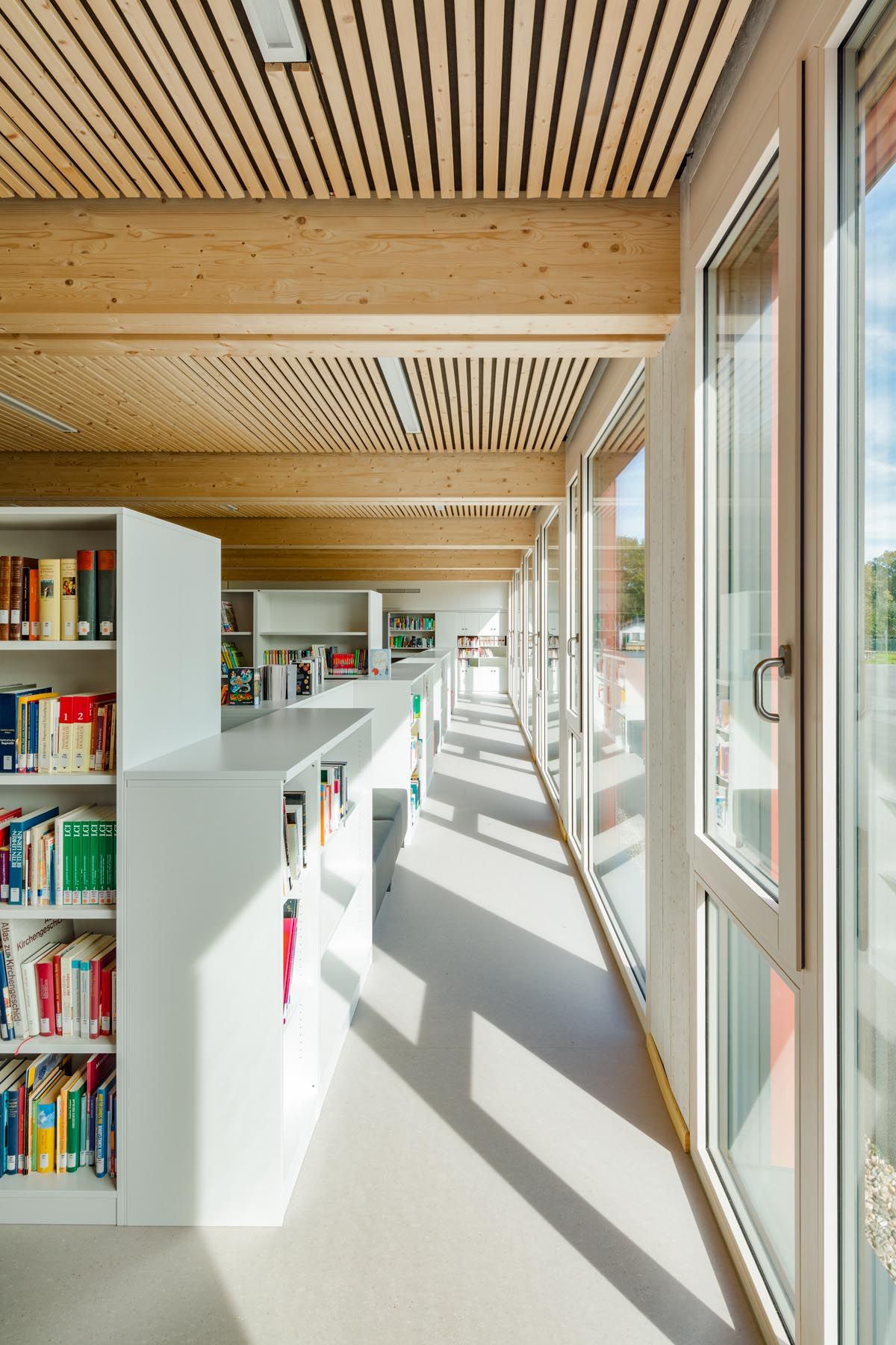 Schulbibliothek mit weißen Regalen entlang bodentiefer Fensterfront