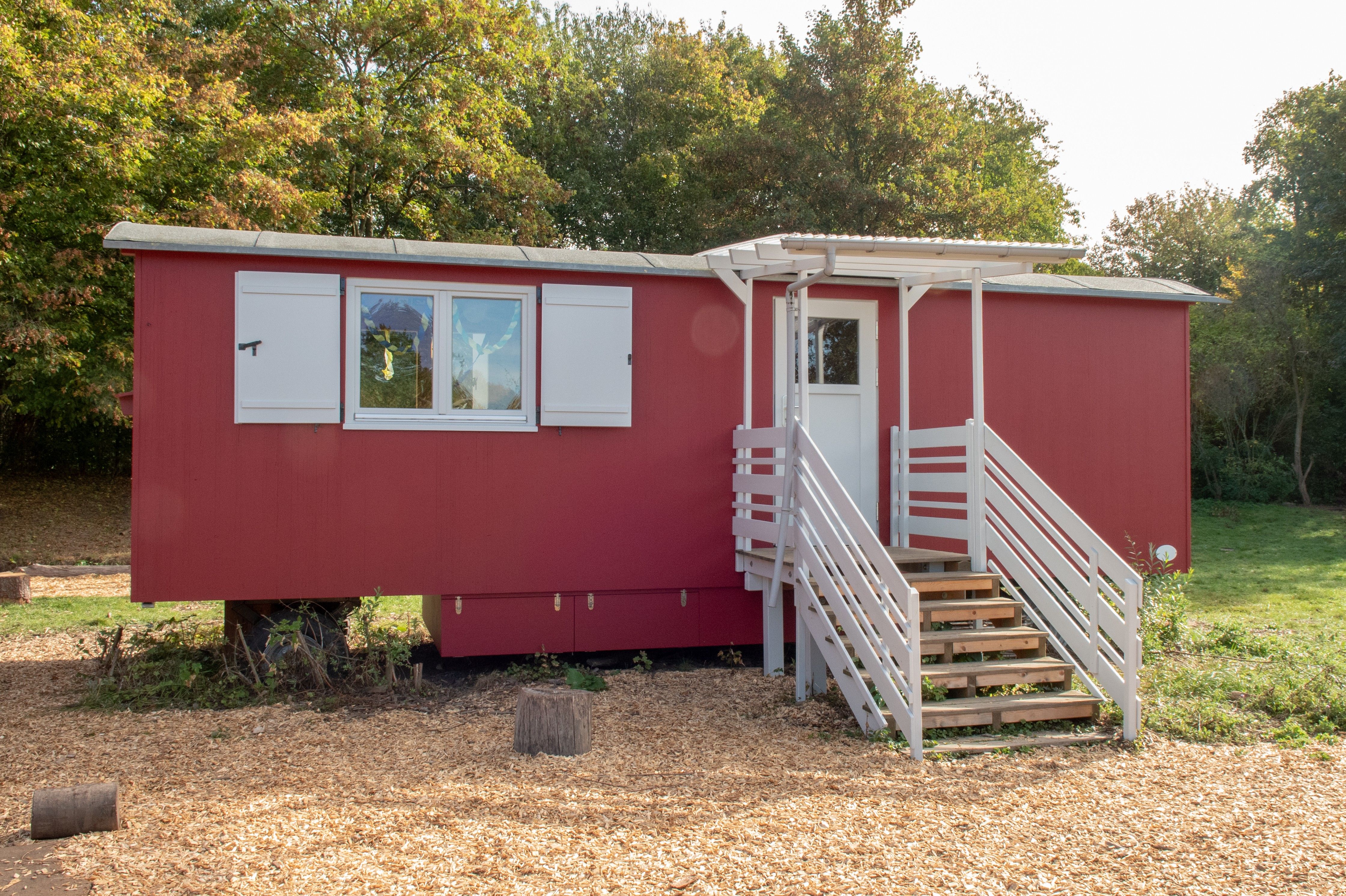 Roter Waldkindergartenwagen mit weißen Fensterläden, weißer Treppe und überdachtem Eingang am Rand einer Waldlichtung