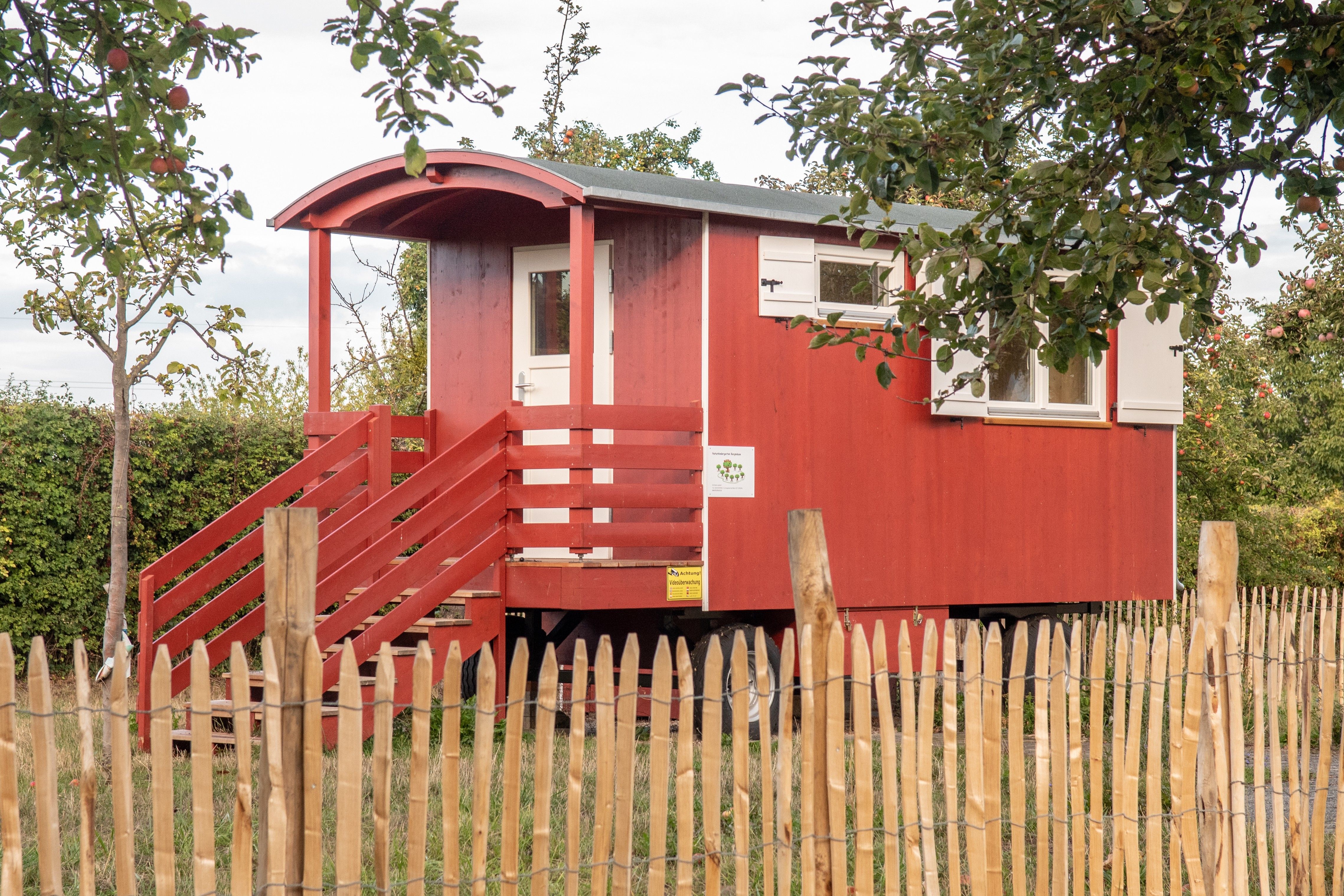 Roter Waldkindergartenwagen mit überdachtem Eingang hinter einem Staketenzaun unter Apfelbäumen