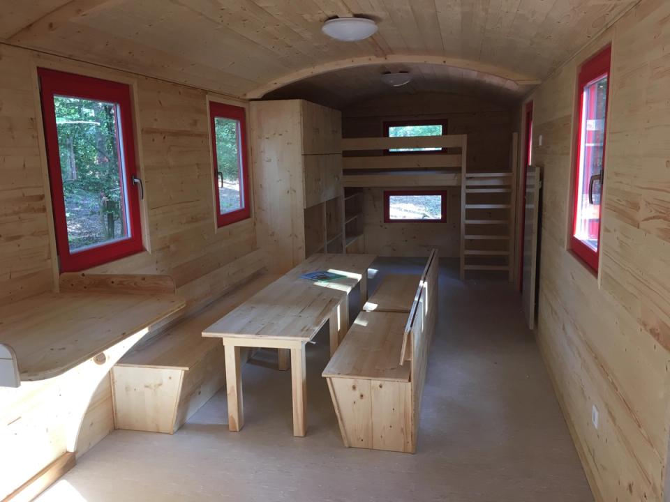 Innenraum mit langen Tischen und Bänken, roten Fensterrahmen und einem Hochbett mit Leiter am Ende