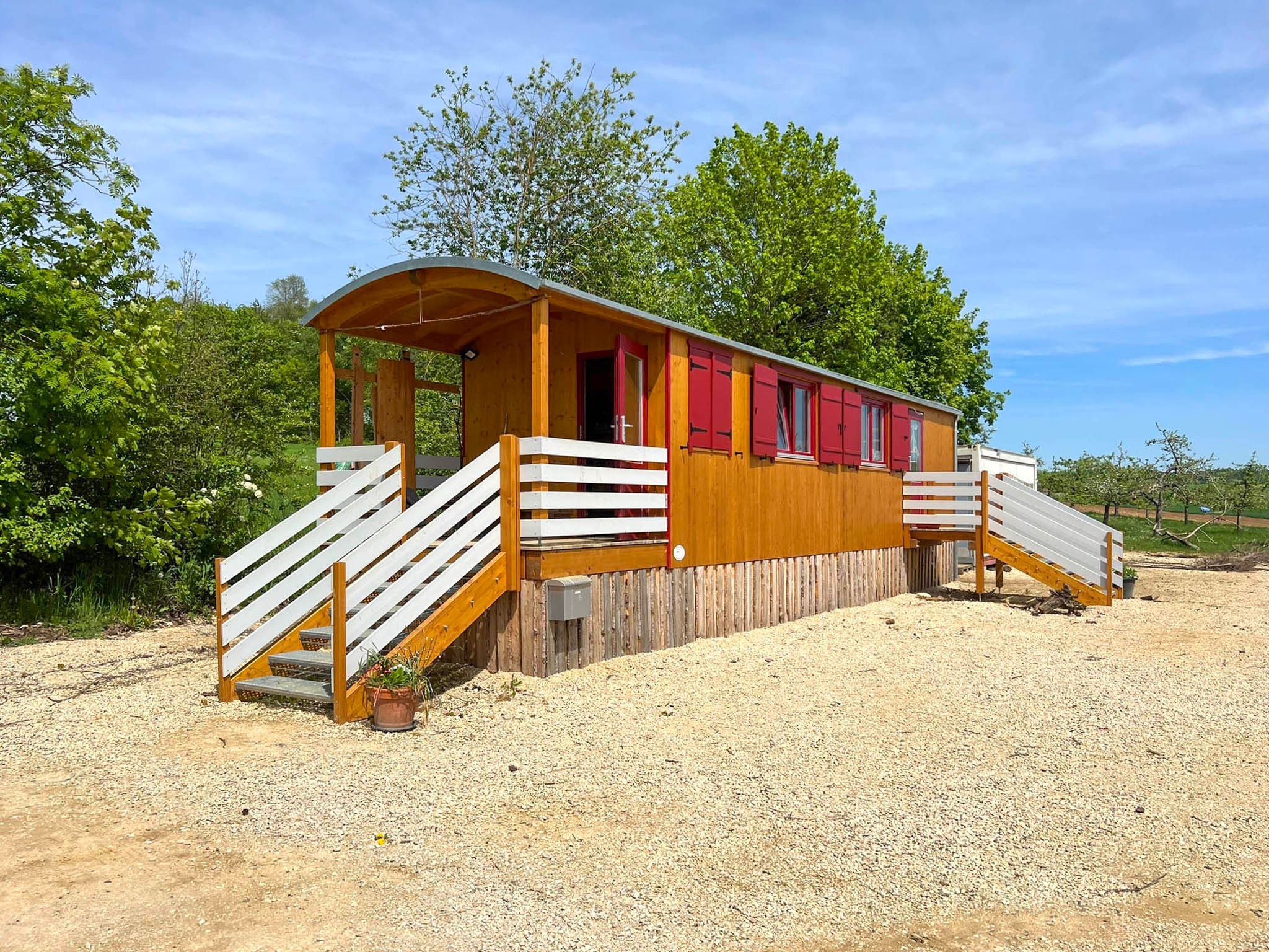 Waldkindergartenwagen aus hellem Holz mit roten Fensterläden und weißer Treppe auf einem Kiesplatz