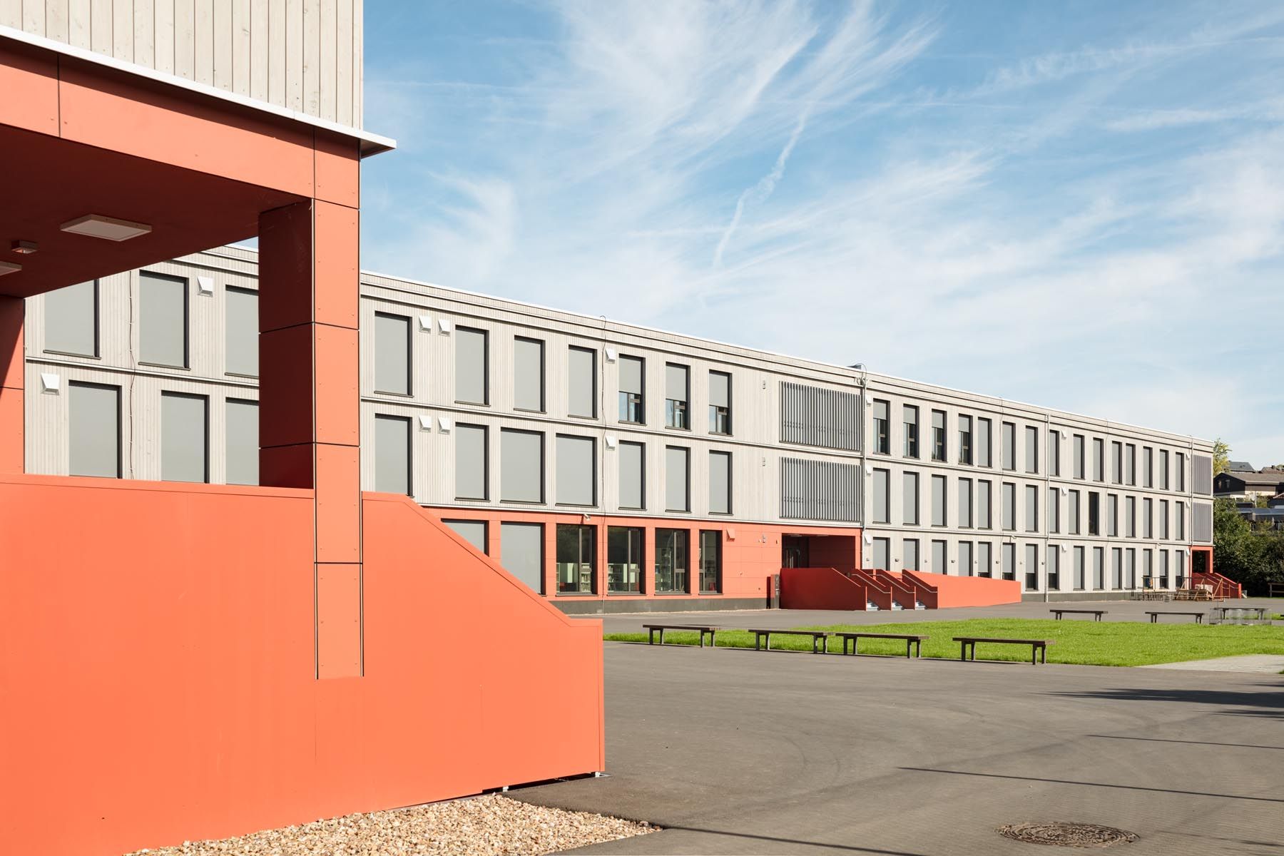 Schulhof der Interimsschule mit rotem Eingang und Bänken vor der hellen Fassade