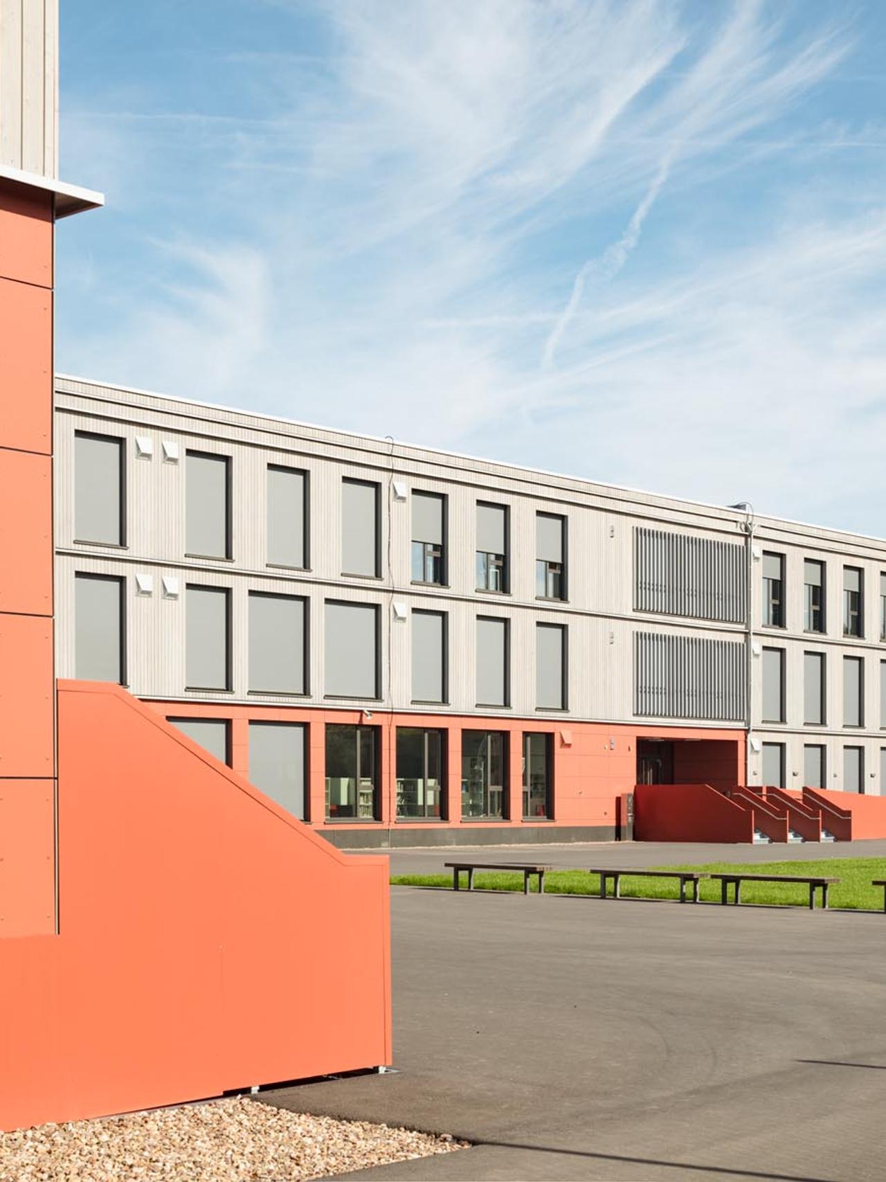 Schulhof der Interimsschule mit rotem Eingang und Bänken vor der hellen Fassade