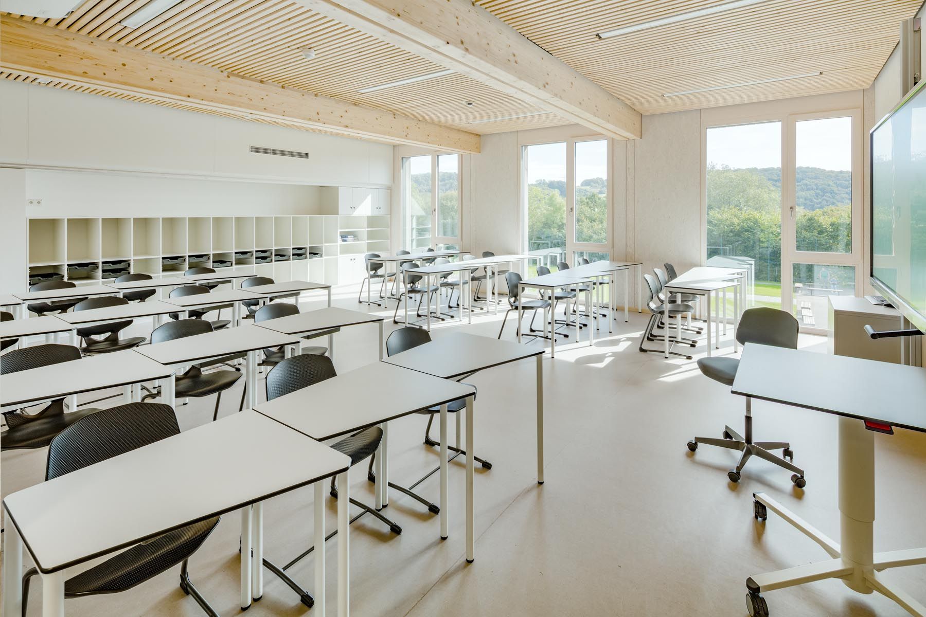 Heller Klassenraum mit Tischreihen, Stauraumwand und raumhohen Fenstern