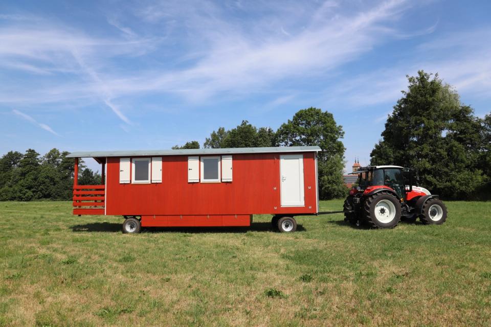 Traktor zieht einen roten Waldkindergartenwagen über eine Wiese, darüber ein weiter Himmel mit Schleierwolken
