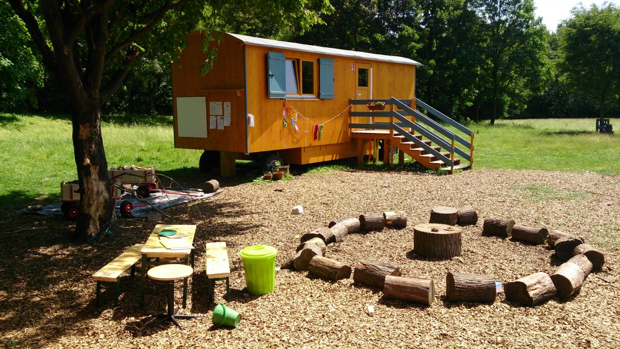 Waldkindergartenwagen mit blauen Fensterläden auf einer Lichtung, davor ein Sitzkreis aus Baumstämmen