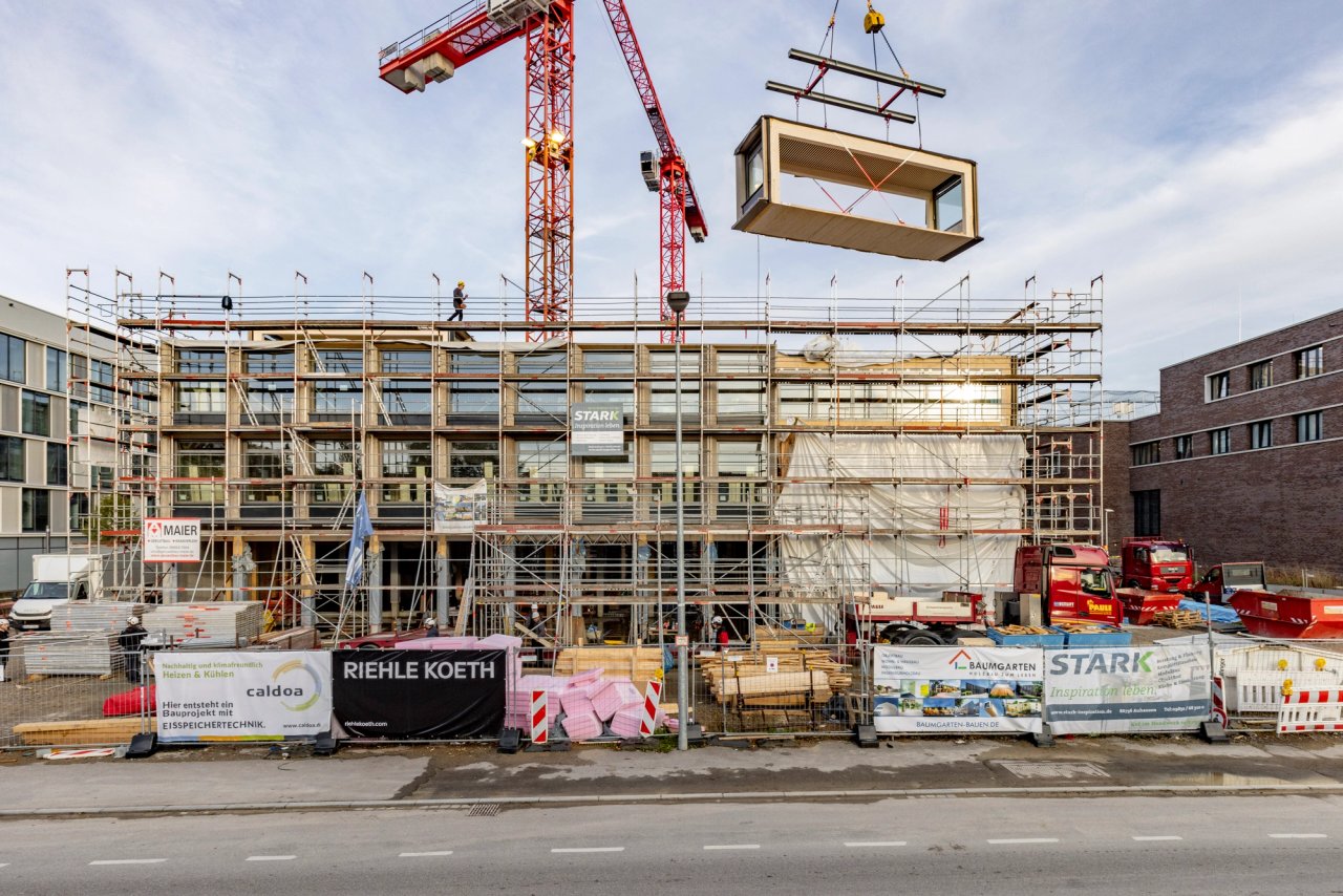 Holzmodul schwebt am Abend am Kran über dem eingerüsteten Neubau in Stuttgart
