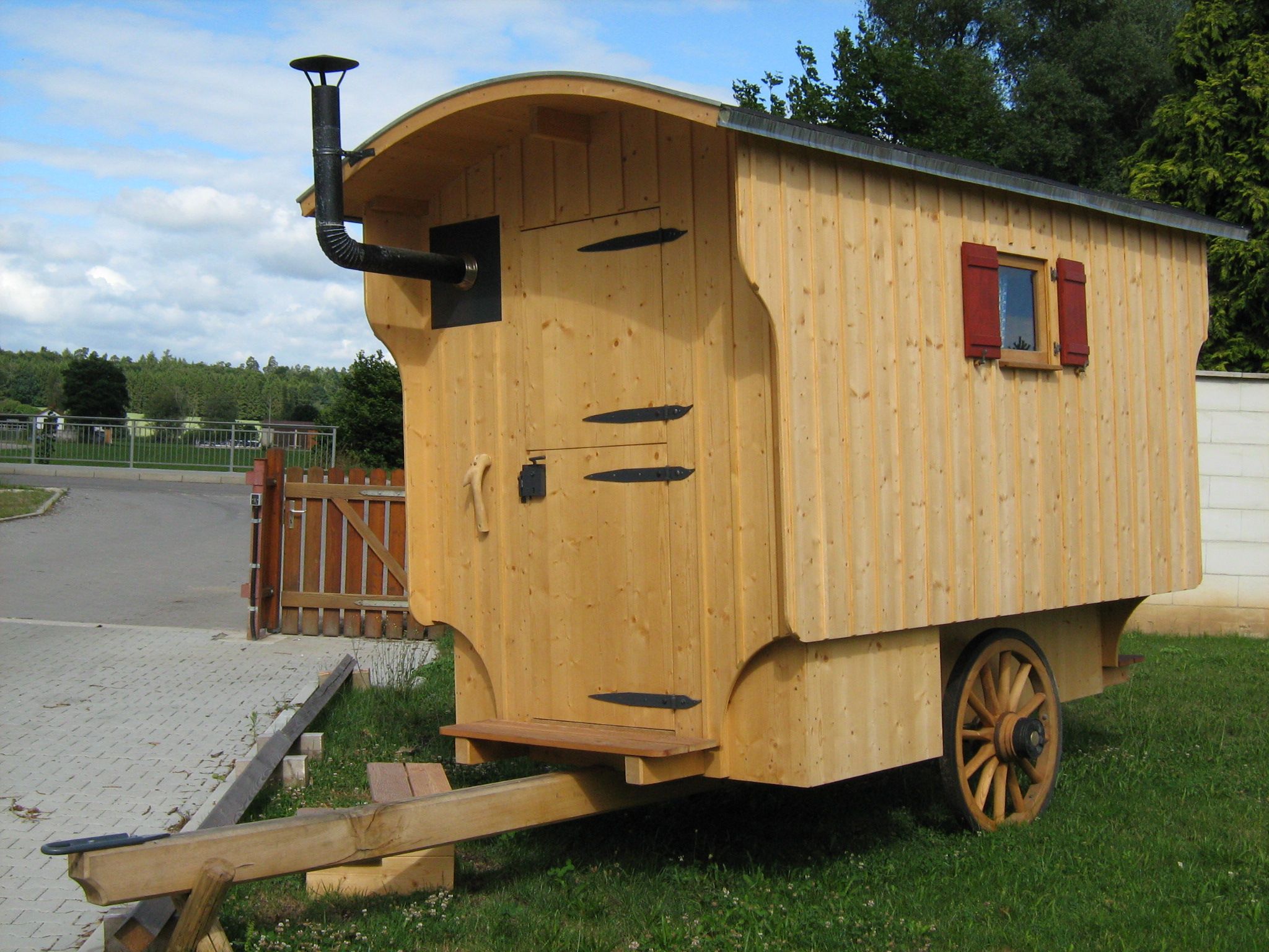 Gartenwagen aus hellem Holz mit roten Fensterläden, Ofenrohr und Speichenrad