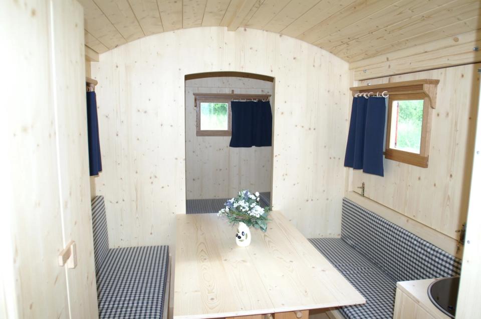 Innenraum eines Schäferwagens mit Tisch, gepolsterten Bänken und blauen Vorhängen