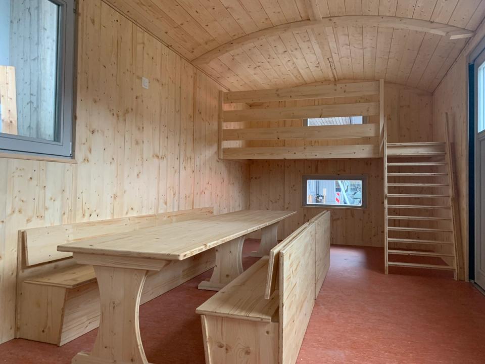 Innenraum mit langen Tischen und Bänken, hinten eine Hochebene mit Leiter