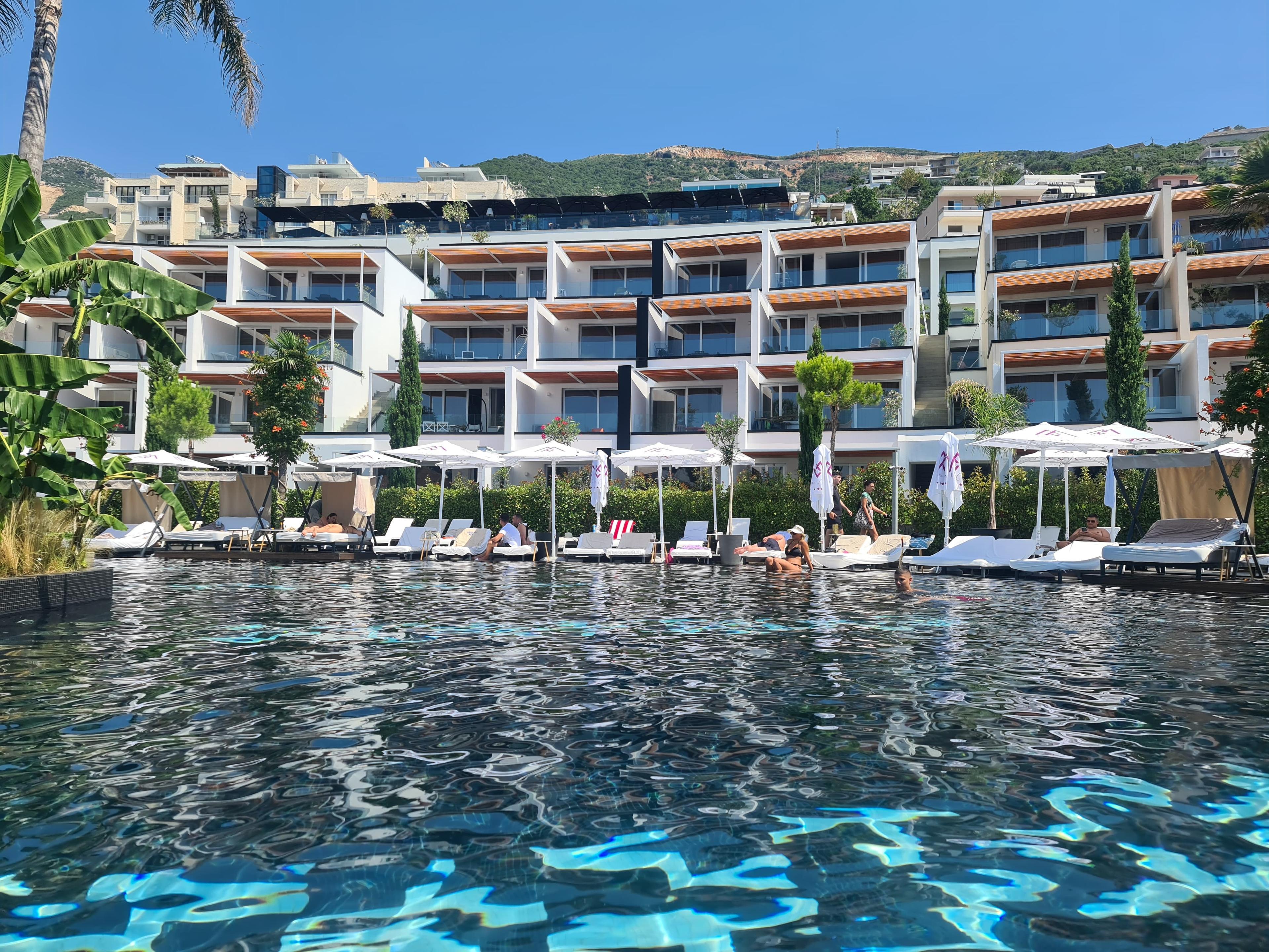 Complesso di appartamenti di lusso con piscina a sfioro e area relax sul mare a Valona, Albania. Nella foto si vedono vegetazione tropicale, lettini con ombrelloni e vista sul mare blu e isole vicine.