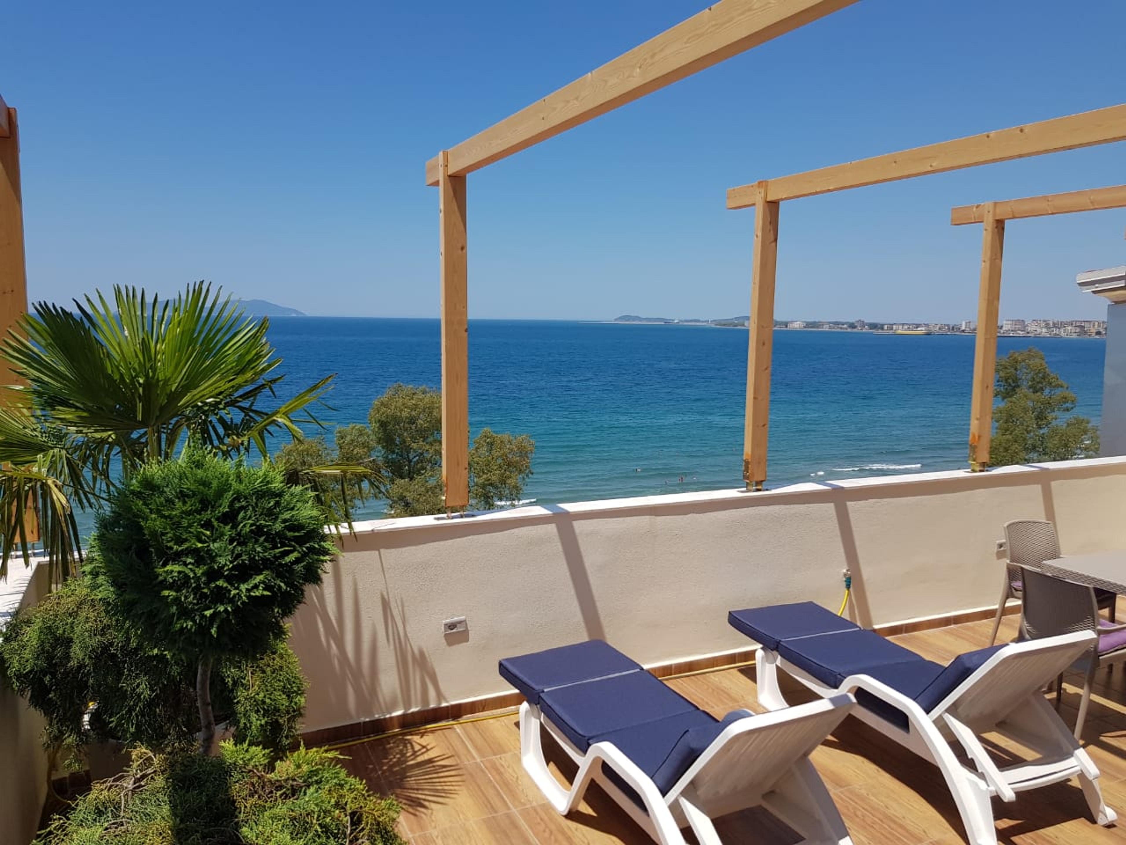 Moderni terassi myytävänä olevassa huoneistossa Vlora, Albania, jossa on panoraamanäköala merelle. Terassilla on mukavia aurinkotuoleja, pöytä ja koristekasveja, ja taustalla näkyy hiekkaranta ja Vloran rannikko kirkkaan taivaan alla.