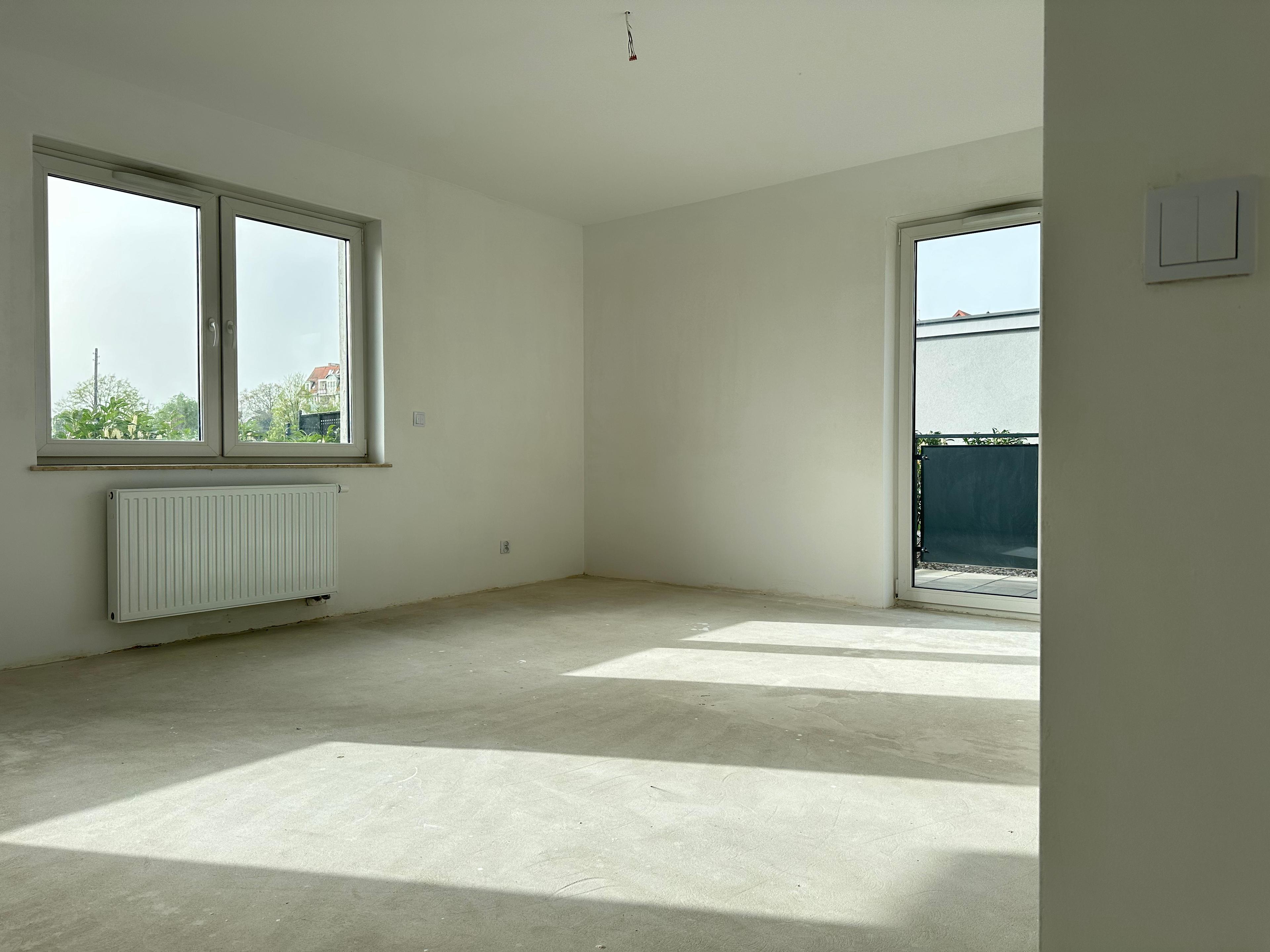 Chambre spacieuse dans un nouvel appartement en état de développement – grande fenêtre et portes-fenêtres vitrées offrant un accès à la lumière naturelle. Intérieur lumineux avec radiateur sous la fenêtre et espace ouvert pour aménagement personnel.