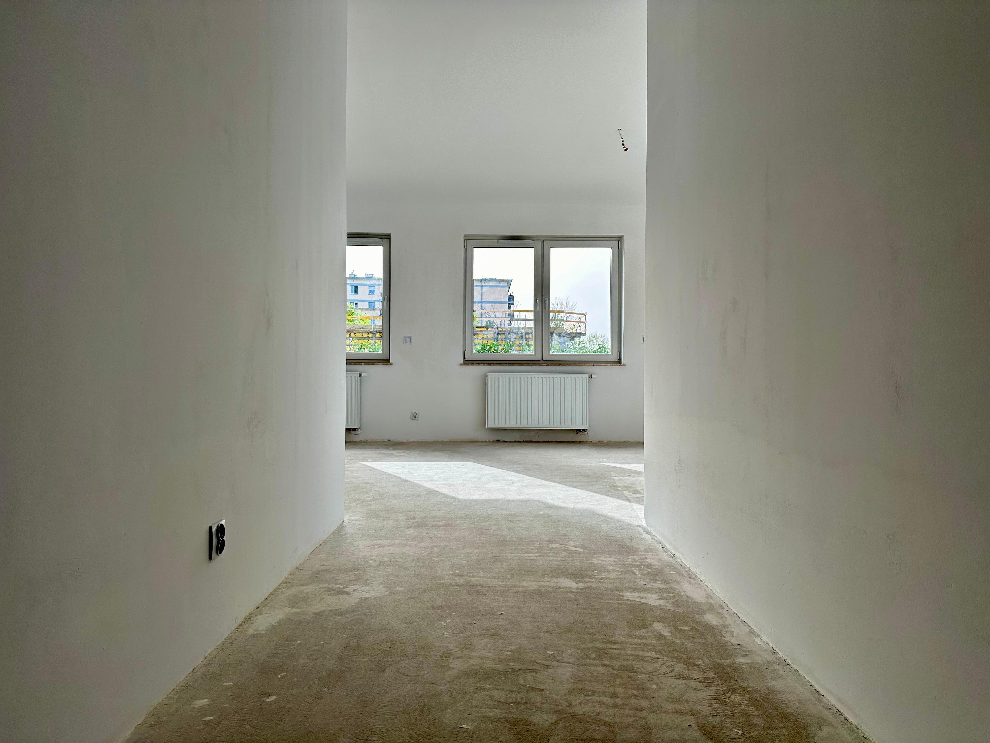 Appartement moderne en état de développement – couloir lumineux menant au salon spacieux avec trois fenêtres et un radiateur. Lumière naturelle pénétrant à l'intérieur soulignant un design minimaliste. Espace idéal pour l'aménagement.