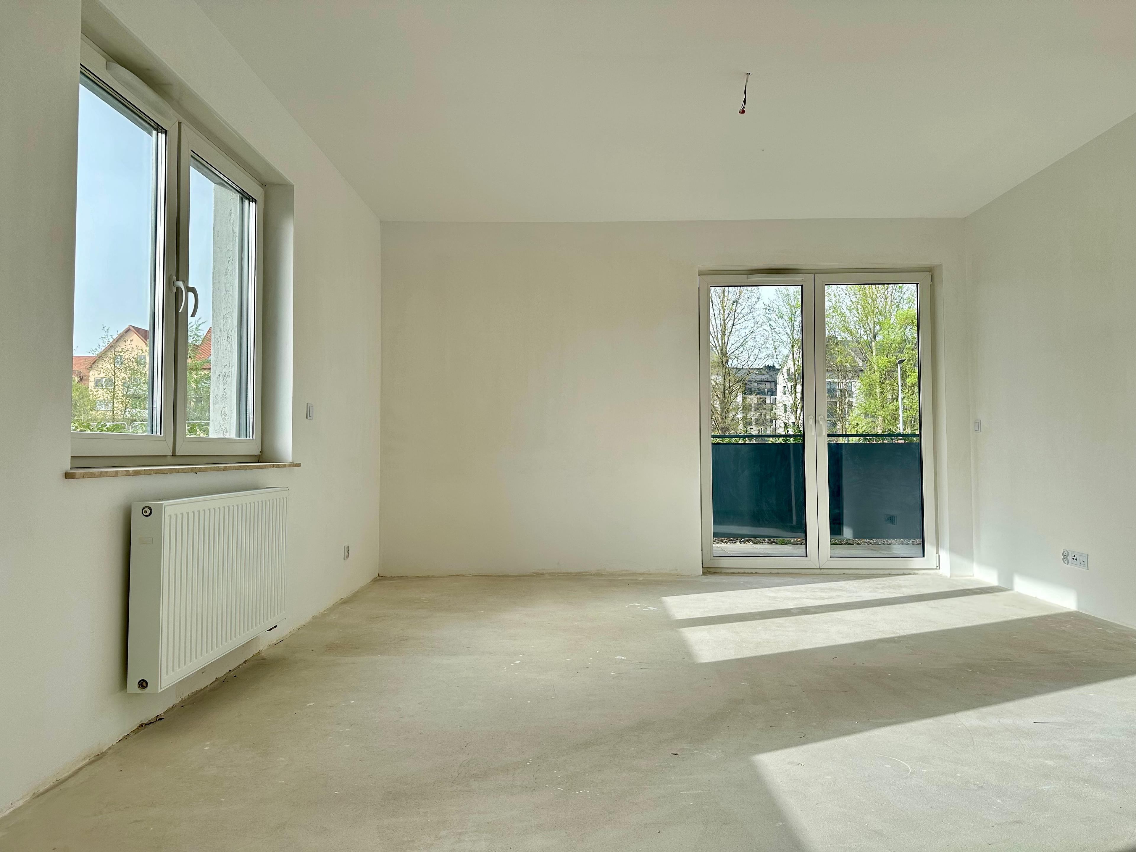 Appartement moderne en état de développement – salon lumineux avec grande fenêtre et sortie vitrée sur le balcon. Intérieur spacieux avec murs blancs, radiateur et éclairage naturel. Espace idéal pour l'aménagement.